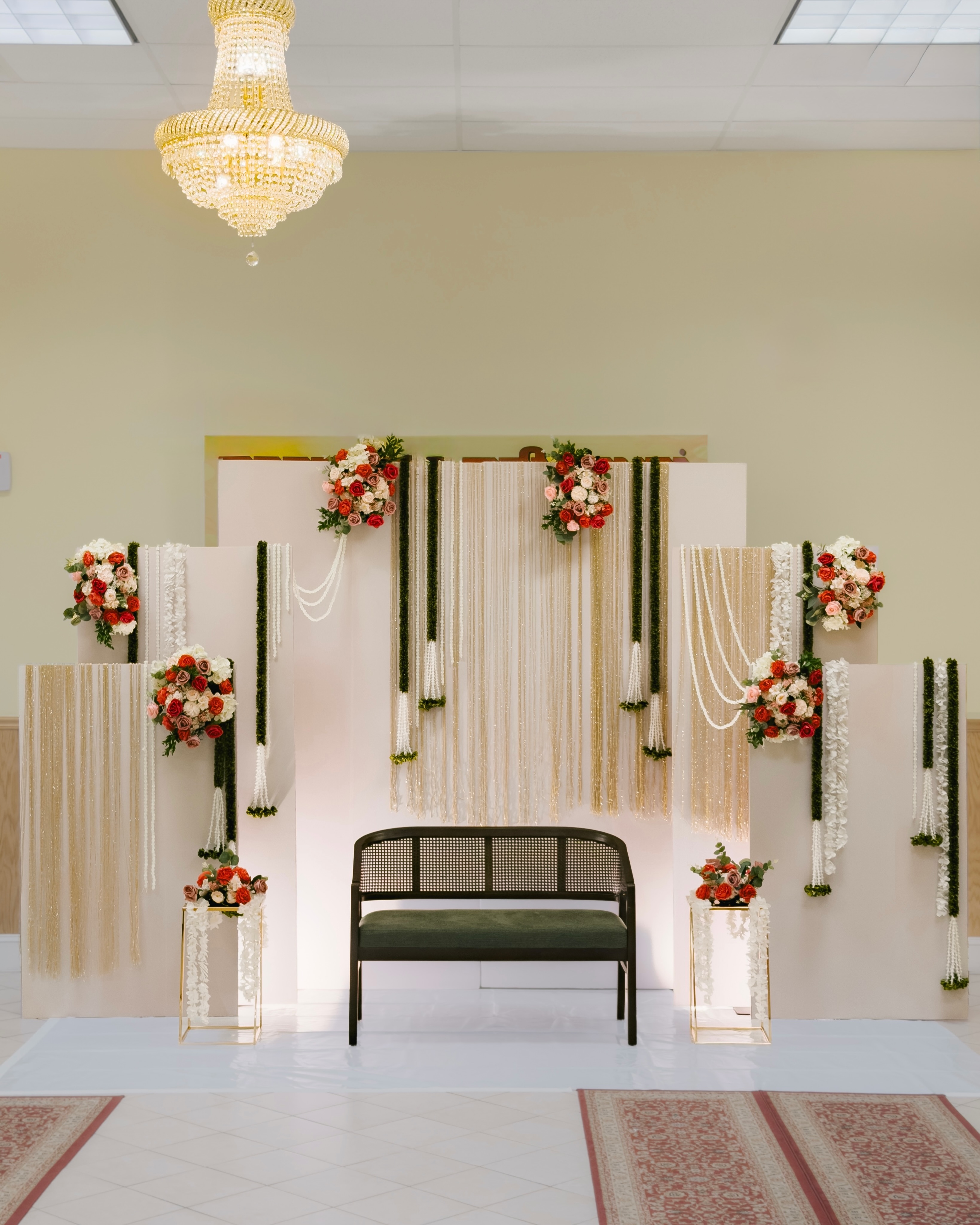 Cream wedding stage with red and white flowers, gold fringe and a dark green settee